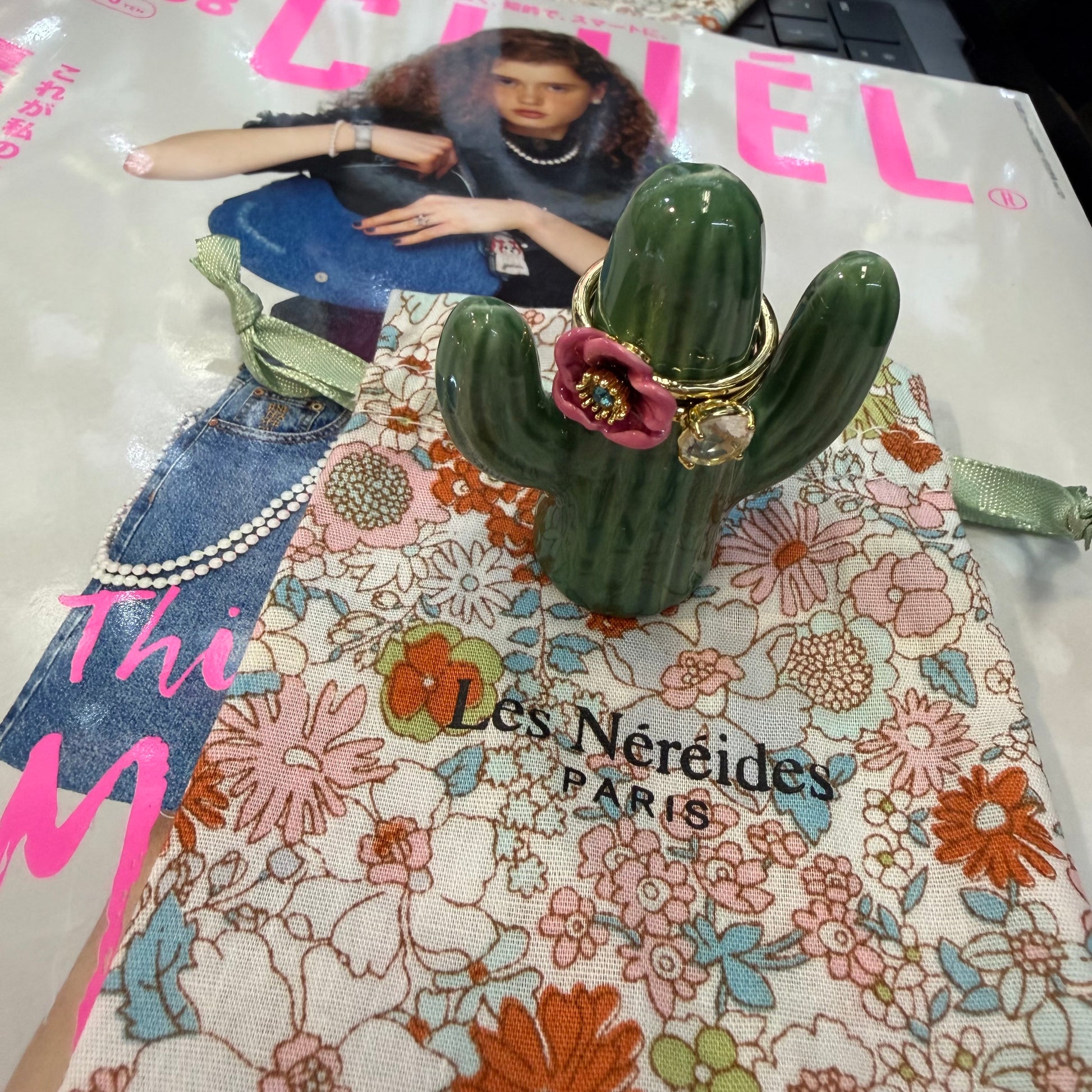 A green cactus-shaped jewelry stand holding two gold rings with a pink flower accent, placed on top of a floral Les Néreides Paris bag. A magazine page with a model is visible in the background.