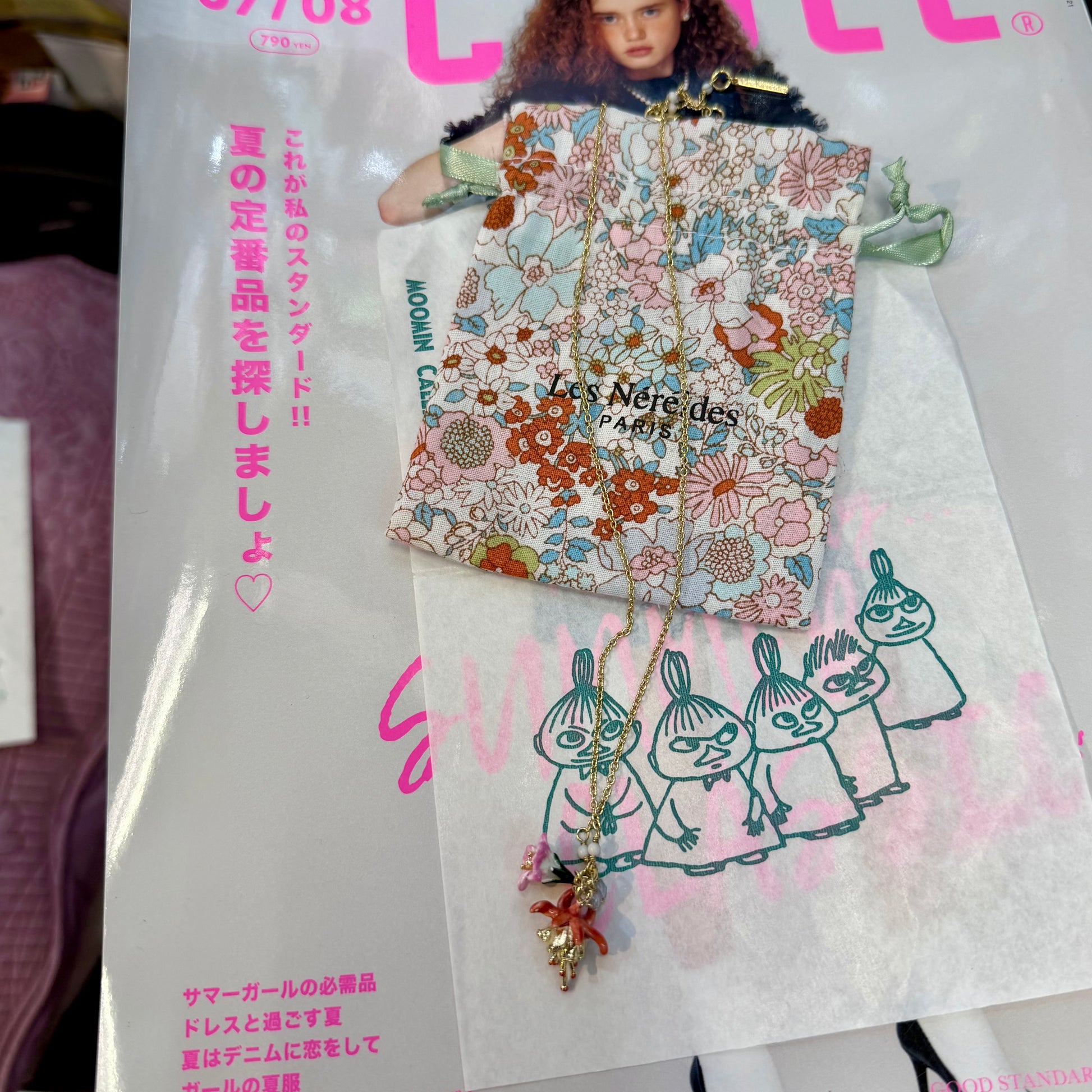 A fashion magazine or booklet featuring a floral-patterned pouch and a gold-tone necklace with a pink and gold pendant, displayed over printed illustrations and pink Japanese text.
